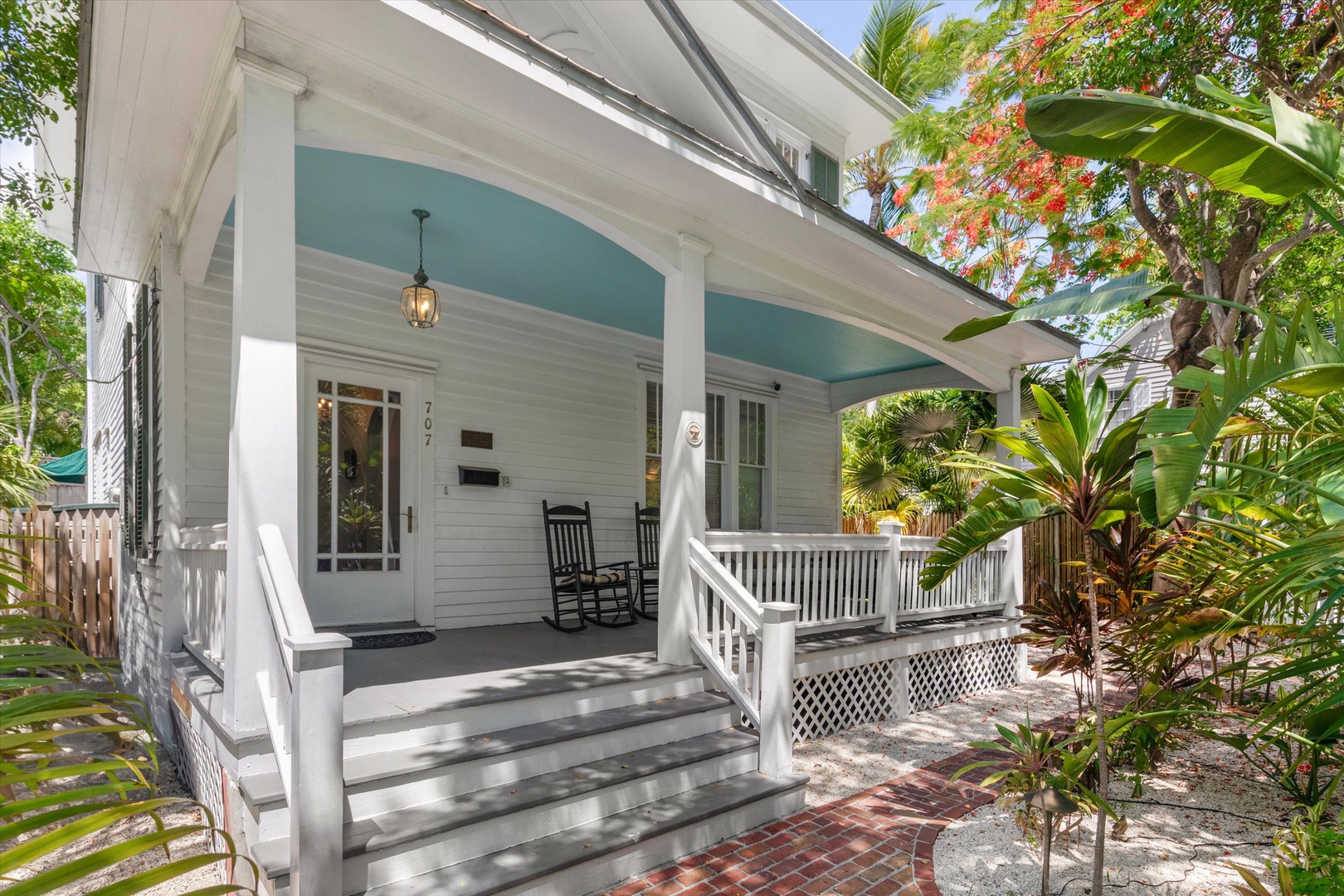 Charming home entrance framed by lush tropical gardens and colorful blooms, offering a peaceful, private arrival experience with classic front porch seating.