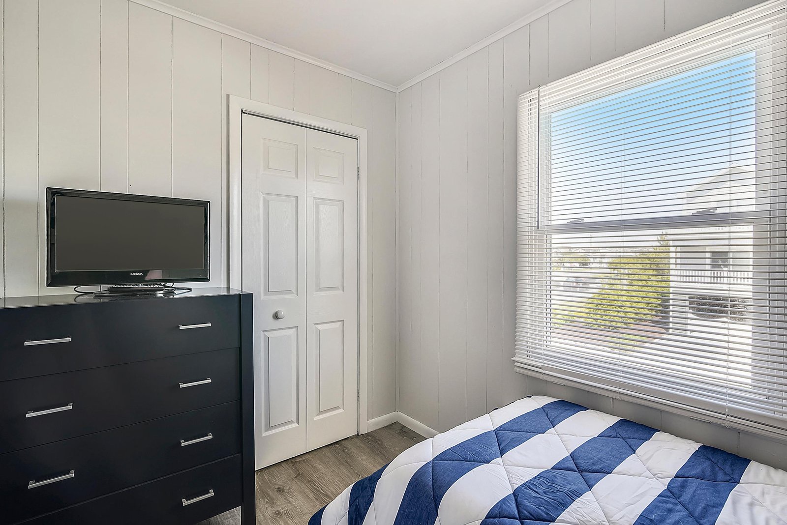 Settle into your cozy bedroom with crisp white paneled walls, natural light streaming through the window, and a comfy bed dressed in navy-and-white striped linens waiting for restful nights.