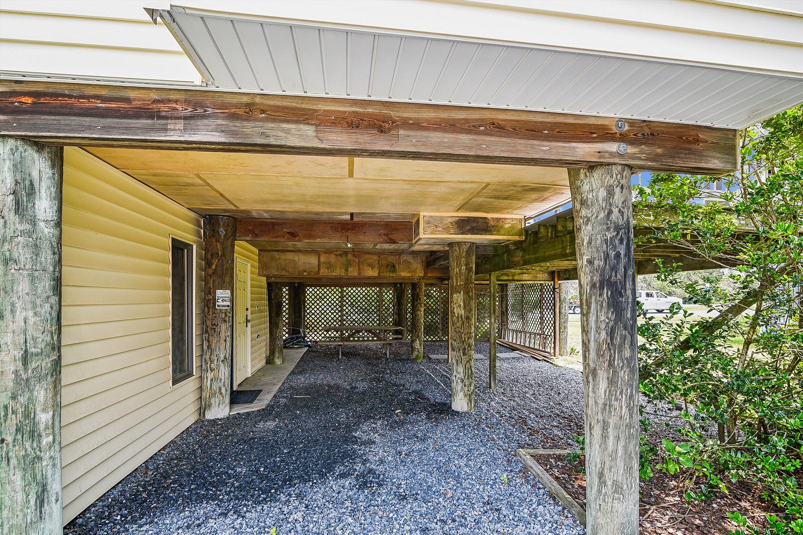 This covered carport provides convenient parking and outdoor protection. The rustic timber structure features gravel flooring and lattice privacy screening, set amid lush greenery.