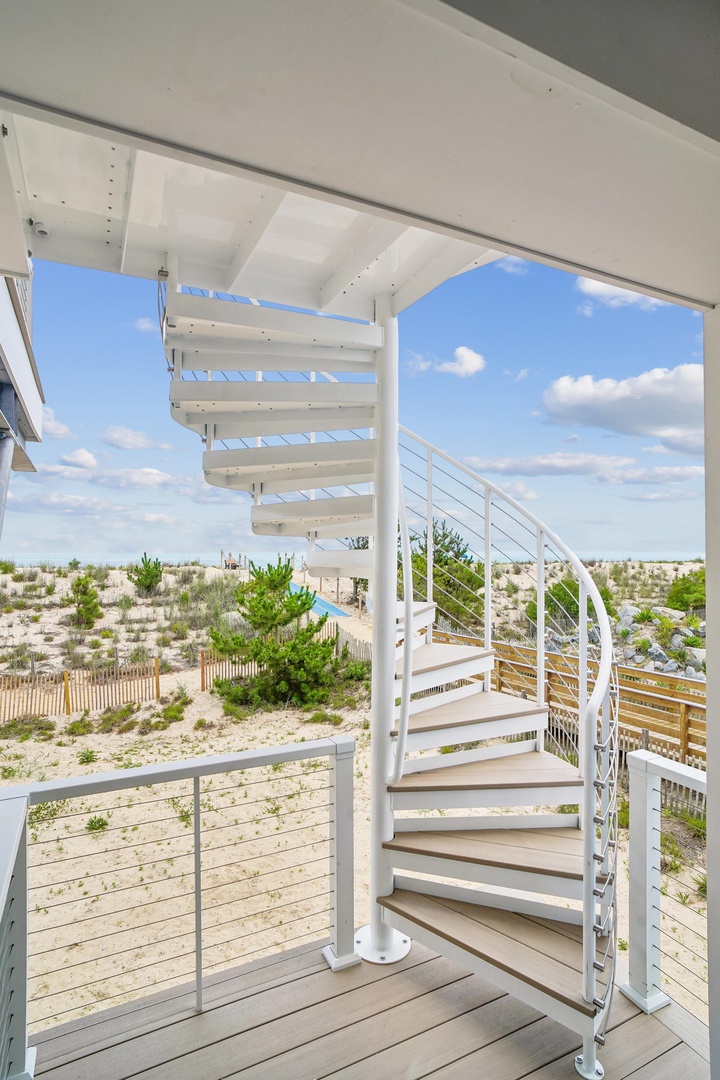 A bright spiral staircase leads from the deck to coastal dunes and clear skies, framing your beachside escape under a sunny blue canopy.