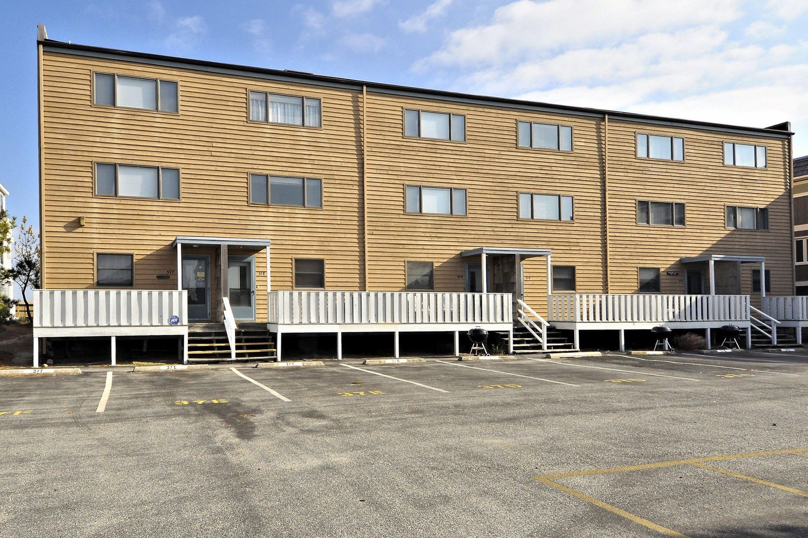 Modern three-story property building with horizontal wood siding and individual unit entrances, featuring parking spaces directly in front.