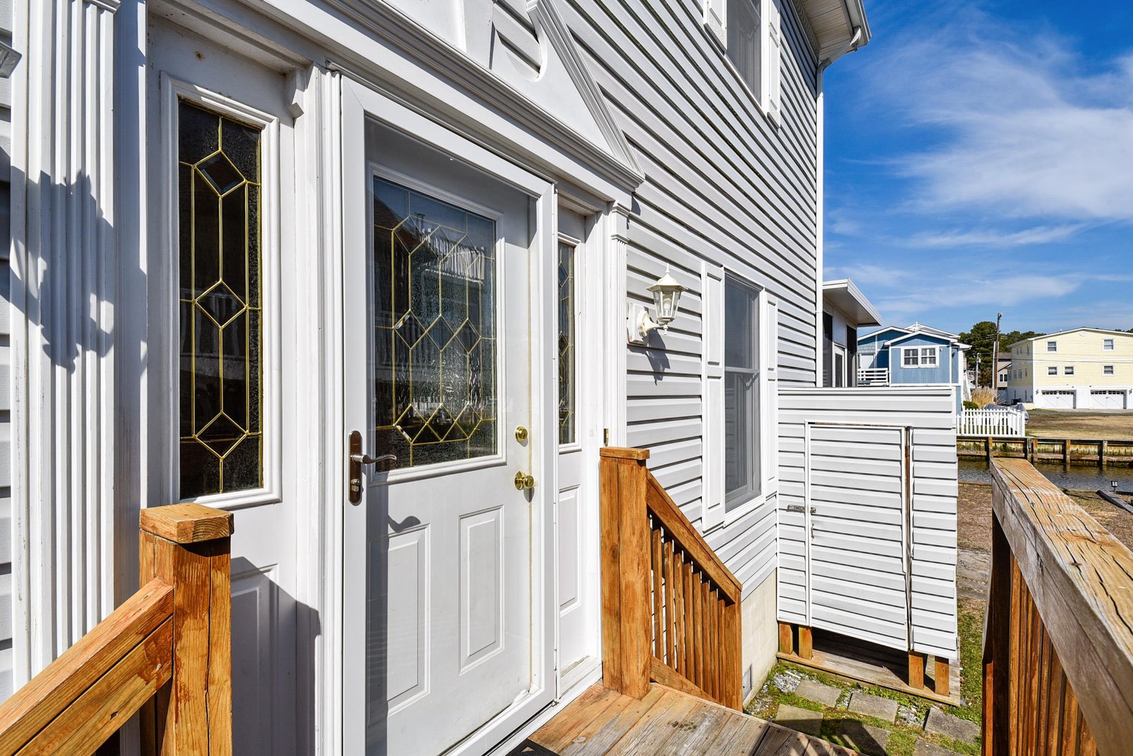 The property features a welcoming entrance with charming decorative glass details and fresh coastal architecture set against a bright sunny day.