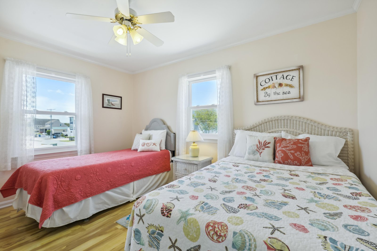 Cozy coastal bedroom with soft morning light from two windows, queen and twin beds dressed in cheerful seashell linens, and a ceiling fan for comfort.