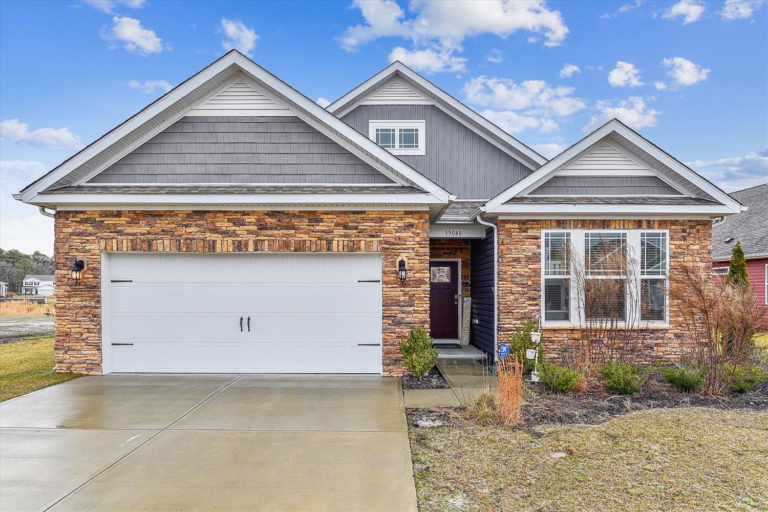 A well-maintained single-story home featuring stone and siding exterior with a two-car garage, surrounded by landscaped beds and open sky views in a quiet residential neighborhood.
