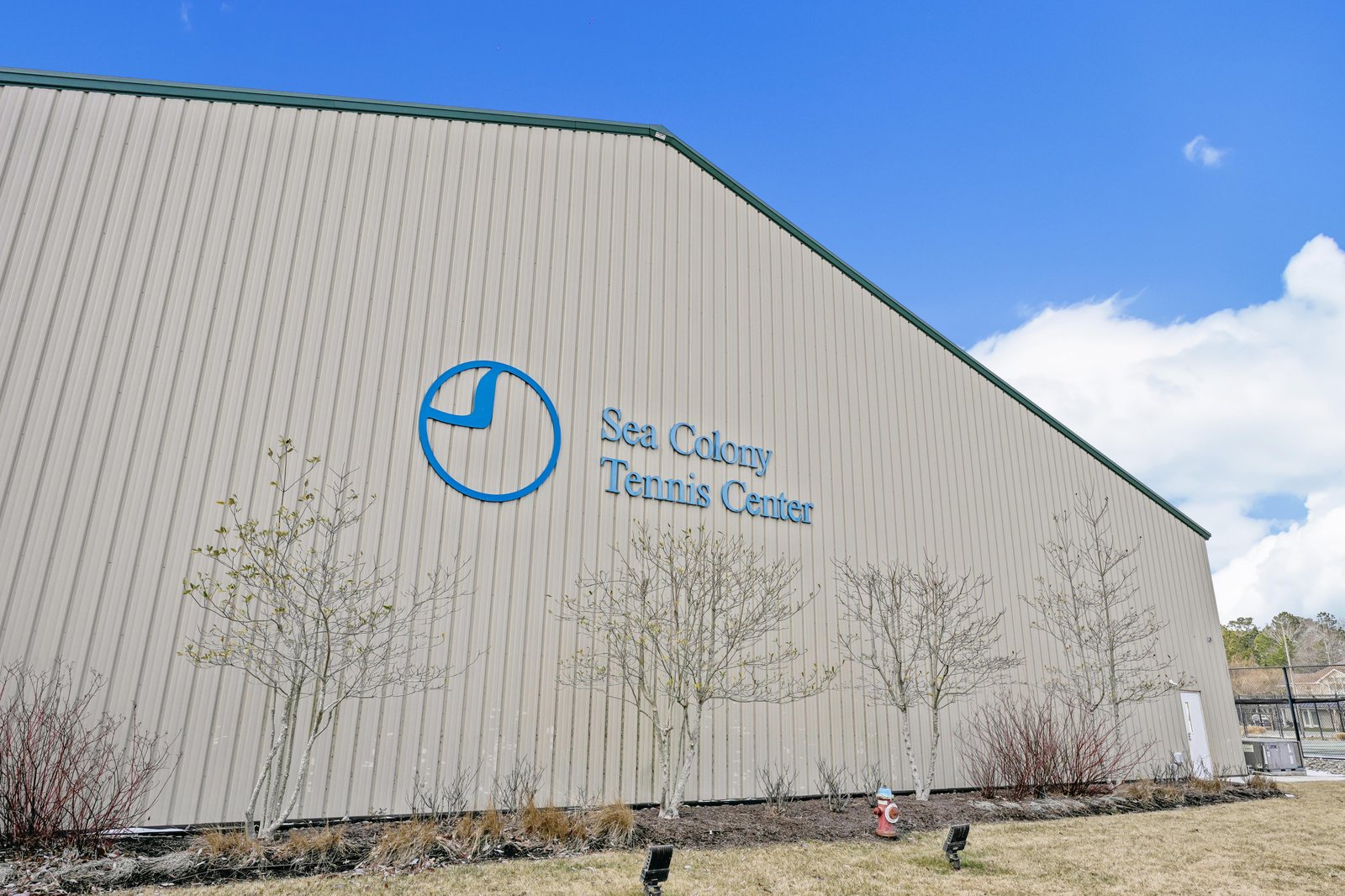The Sea Colony Tennis Center exterior, featuring a distinctive blue logo on the building facade, surrounded by landscaping and welcoming signage.