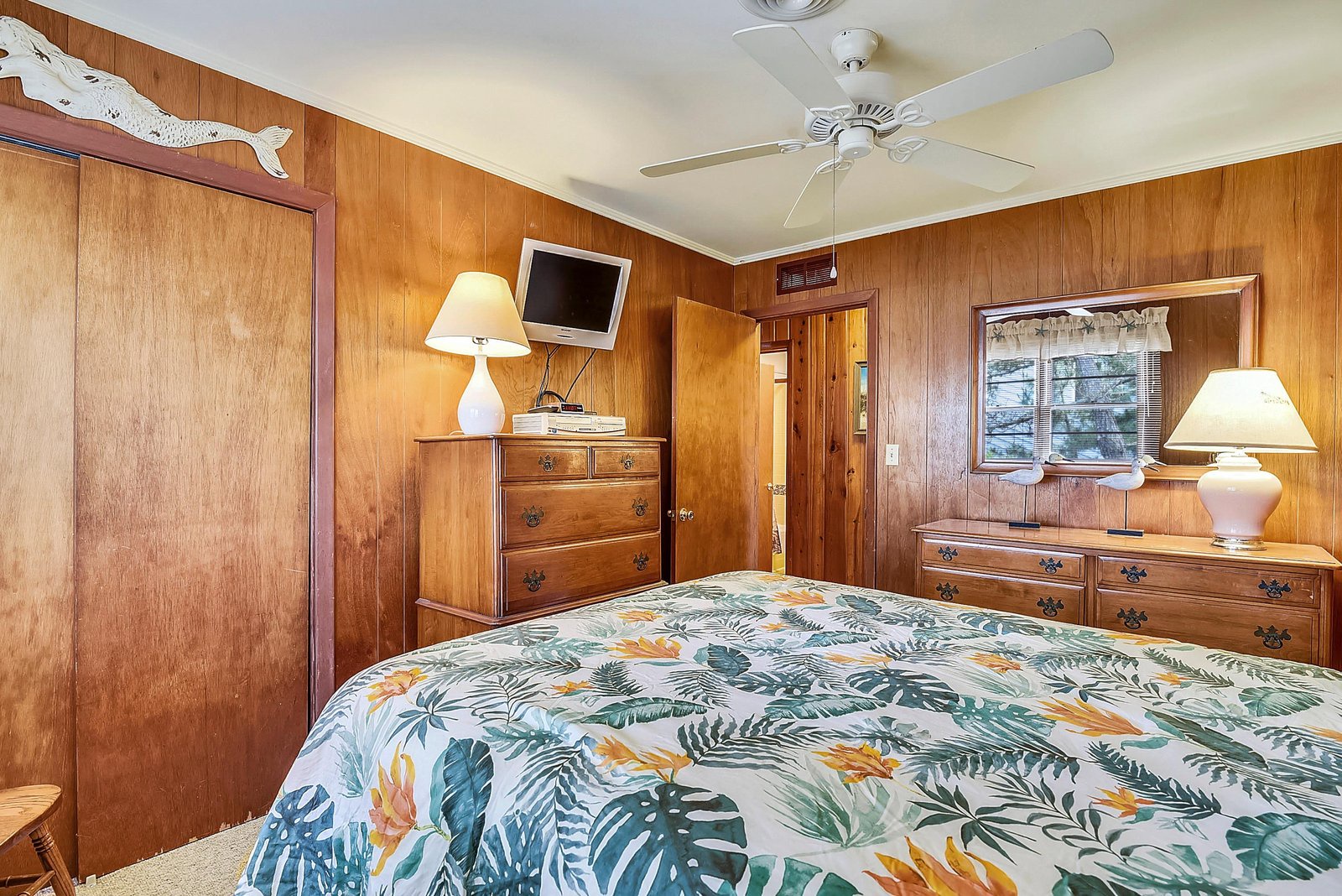 Step into your cozy retreat with warm wood paneling and charming vintage furnishings that create a nostalgic, homey atmosphere. The ceiling fan keeps you comfortable while tropical bedding adds a playful touch.