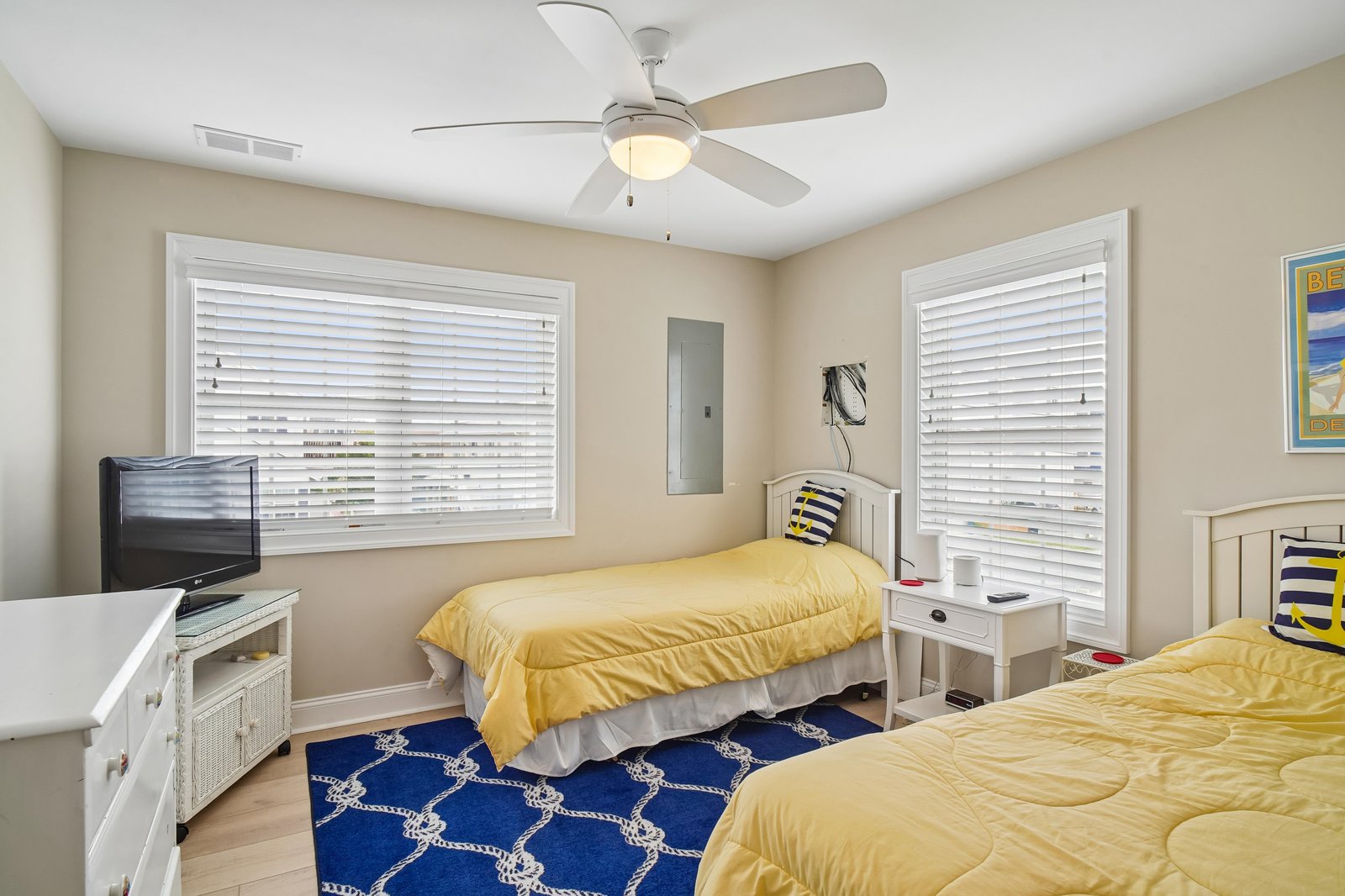 Your bright and cheerful bedroom awaits with twin beds dressed in sunny yellow linens, a nautical-themed rug underfoot, and a gentle ceiling fan to keep you cool. Natural light pours in through large windows, creating a welcoming coastal retreat.