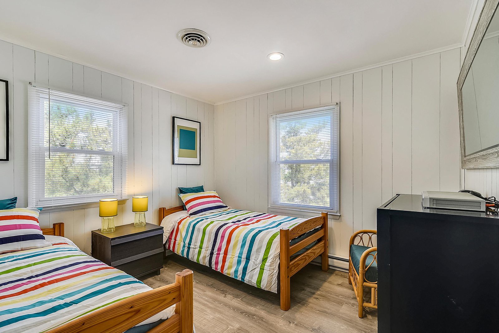 Your bright getaway bedroom features twin beds with cheerful striped linens, warm wood tones, and sunny windows overlooking greenery—perfectly cozy for restful nights and leisurely mornings.