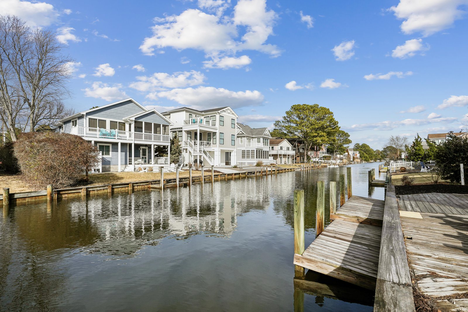 Waterfront property with private dock on calm canal, surrounded by coastal homes and lush greenery under a peaceful blue sky.
