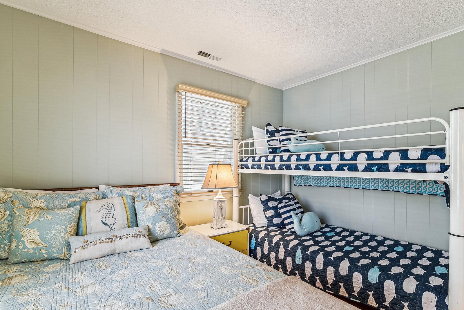 You'll love this charming coastal bedroom with a queen bed and bunk beds, dressed in soothing seafoam tones and ocean-inspired bedding. Perfect for families, it offers comfort and beach-house style in one delightful space.