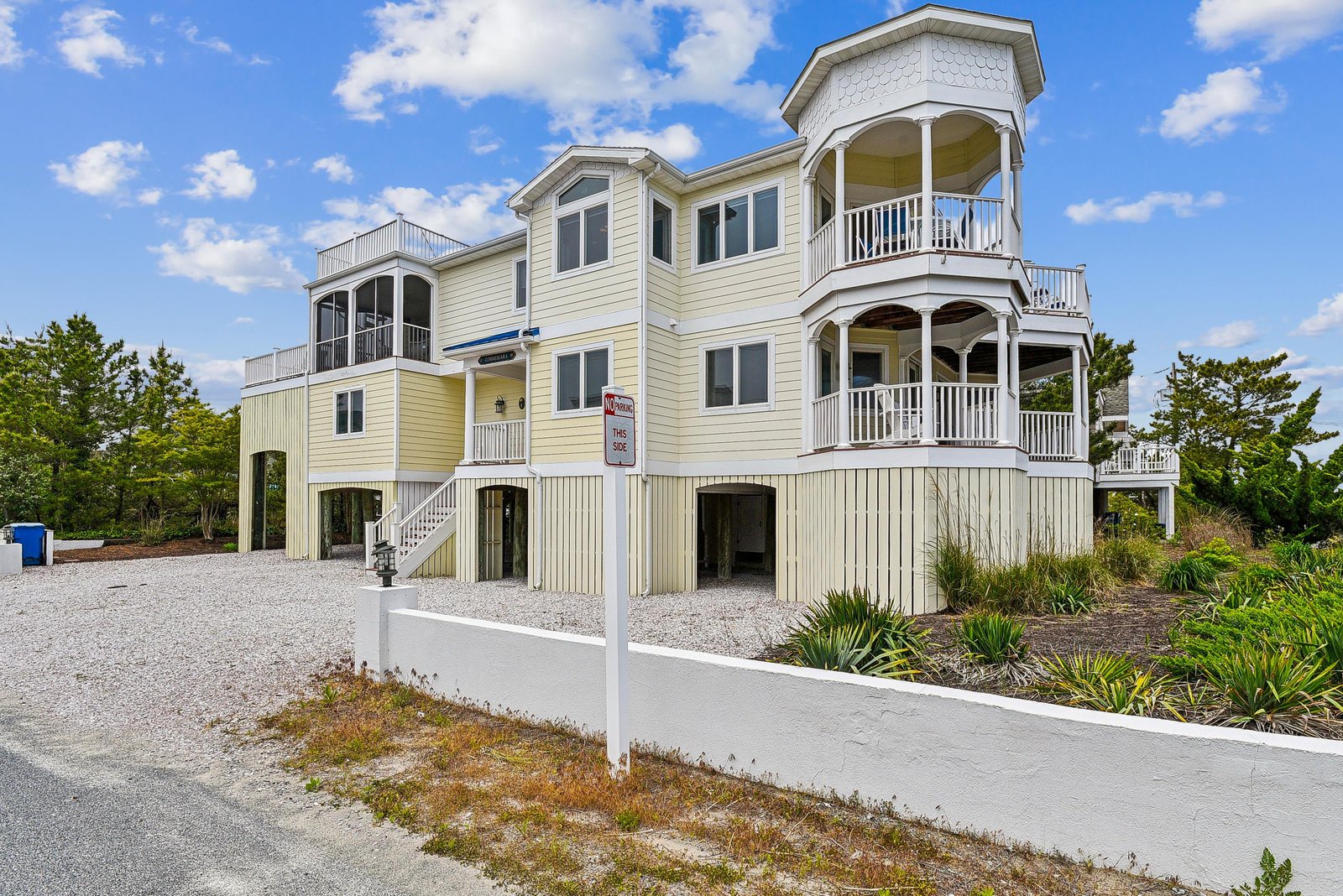 This spacious coastal property features multiple levels of balconies and parking beneath, surrounded by coastal landscaping and set against a sunny blue sky.
