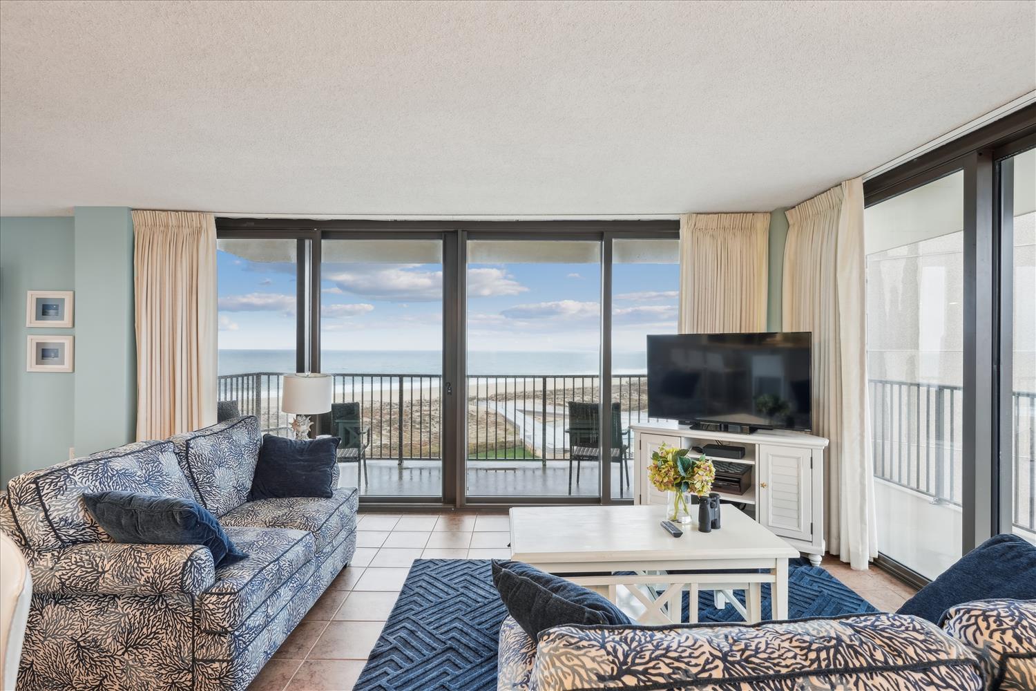 Unwind in your bright living room featuring plush seating, beachy blue accents, and sweeping ocean views framed by floor-to-ceiling glass doors opening to your private balcony.