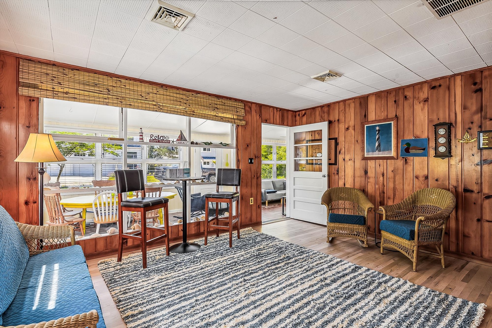 Settle into your cozy coastal living room with warm wood paneling, wicker chairs, and nautical touches. Large windows frame your outdoor dining area, blending comfort with seaside charm.