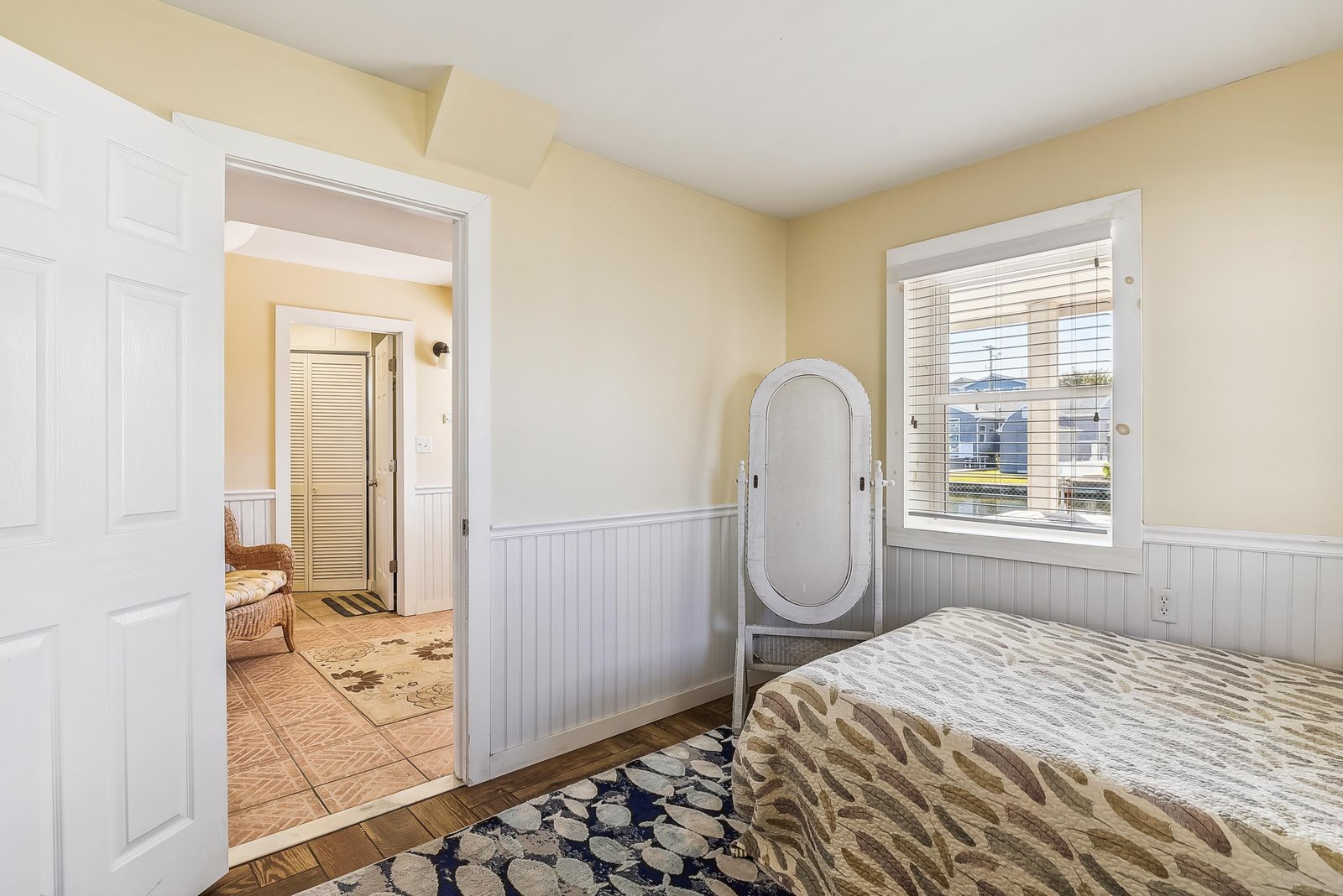 Step into your peaceful bedroom retreat with warm, buttery walls and classic beadboard trim. Natural light pours through the window, while the charming freestanding mirror adds a vintage touch to your coastal getaway.