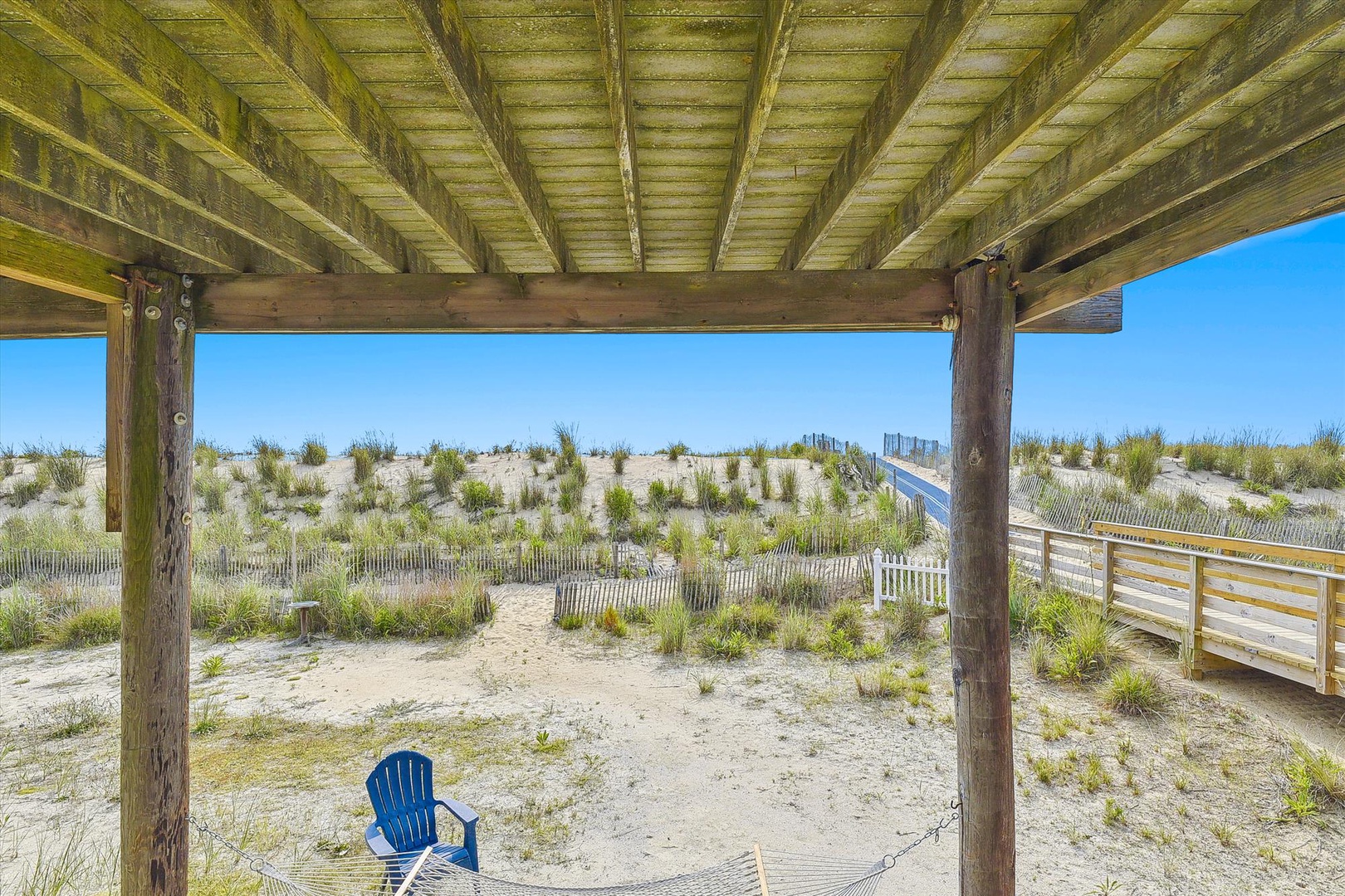 This covered patio provides direct access to the sandy dunes and beach beyond, framed by rustic wooden beams and a bright blue sky. A single chair invites you to relax and take in the coastal breeze.