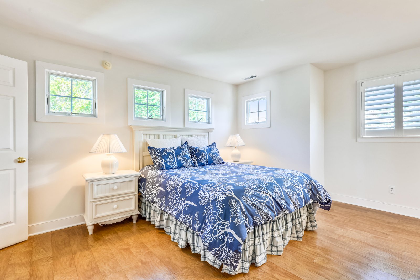 Step into your serene bedroom retreat with crisp white walls, warm hardwood floors, and charming windows framing leafy views. The plush bed awaits with elegant blue linens and soft nightstand lighting.