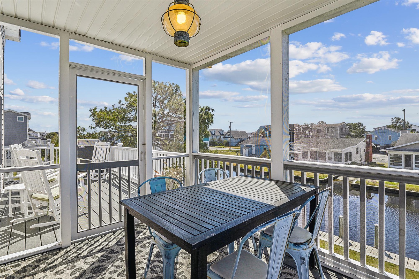 Screened porch with waterfront views over a peaceful residential canal. Dining table for six, nautical pendant light, and surrounding neighborhood scenery create a relaxed coastal setting for the property.