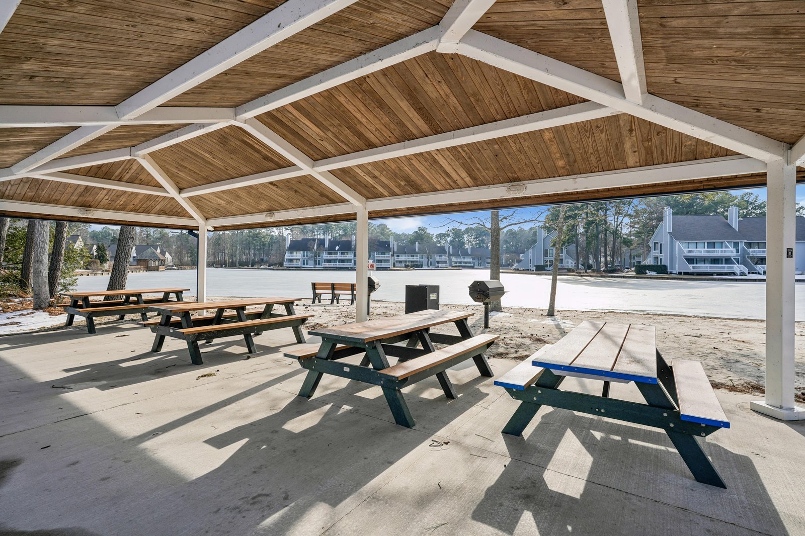 The covered pavilion offers shaded picnic tables and a BBQ grill overlooking the sandy beach and calm lake waters, perfect for outdoor meals and gatherings.