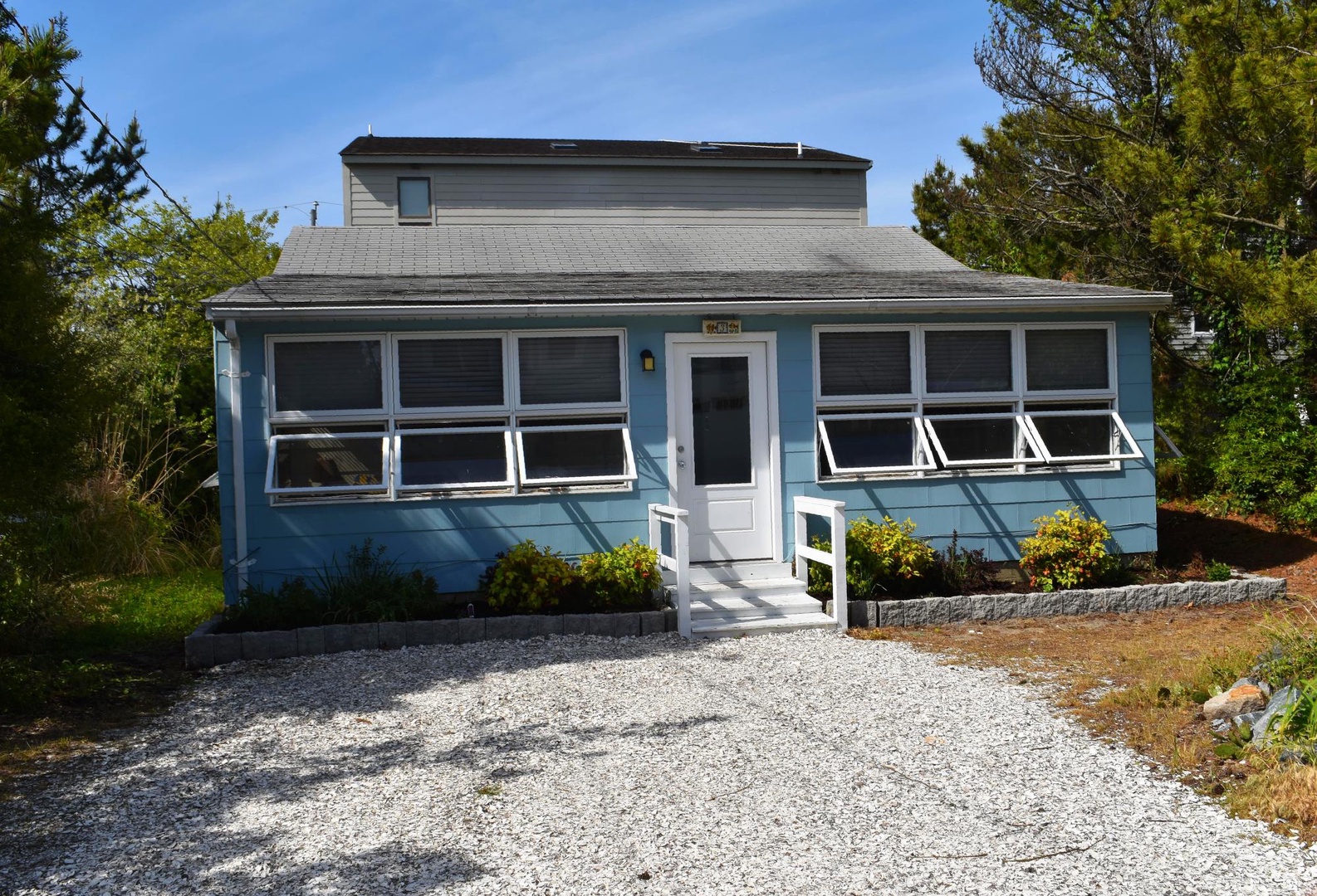 Charming coastal cottage with bright blue exterior, set among mature trees and landscaped grounds. The property features convenient gravel parking and a welcoming front entrance with easy access.