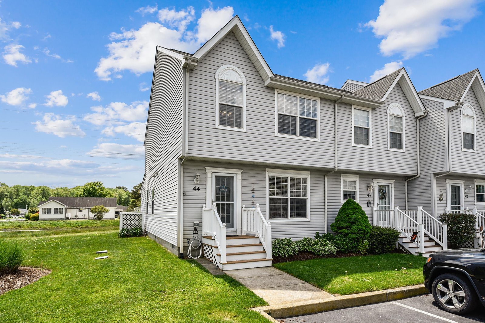 A two-story townhome with classic gray siding, white trim, and a welcoming entrance set in a quiet residential neighborhood with green lawn and mature landscaping.