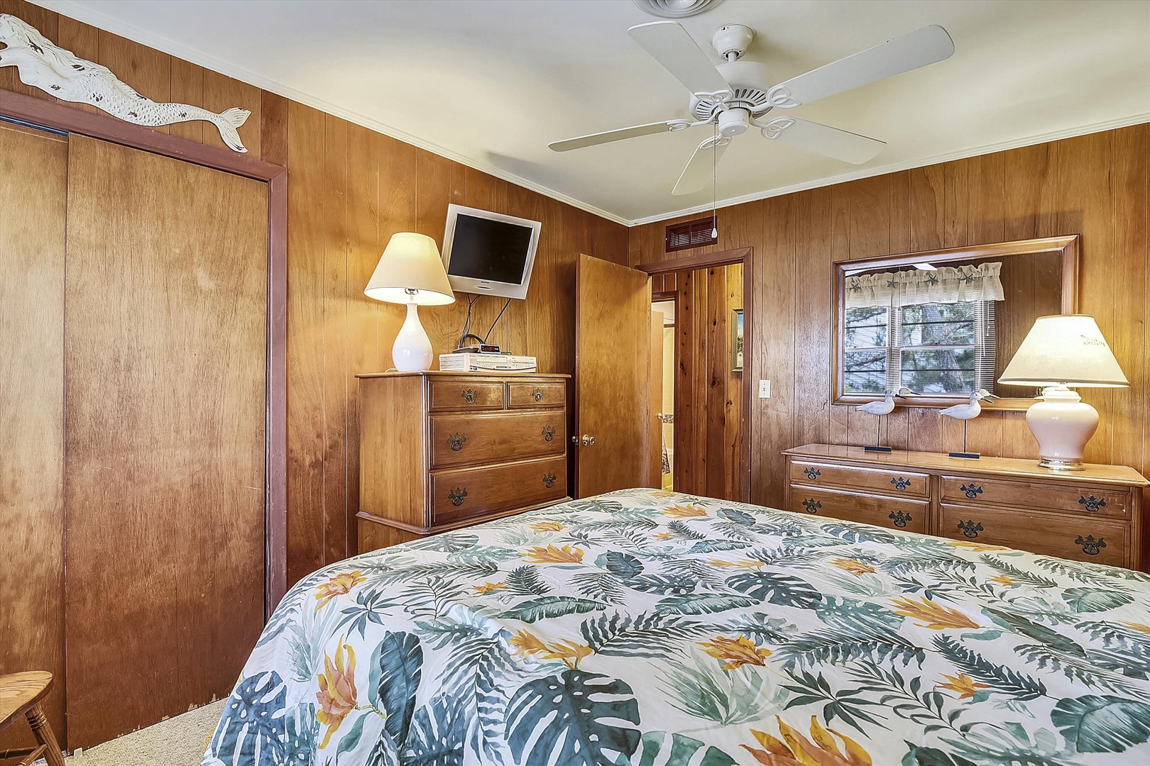 Unwind in your cozy bedroom with warm wood paneling, tropical bedding, and a ceiling fan that keeps you comfortable year-round. Classic furnishings and thoughtful lighting create a peaceful retreat.