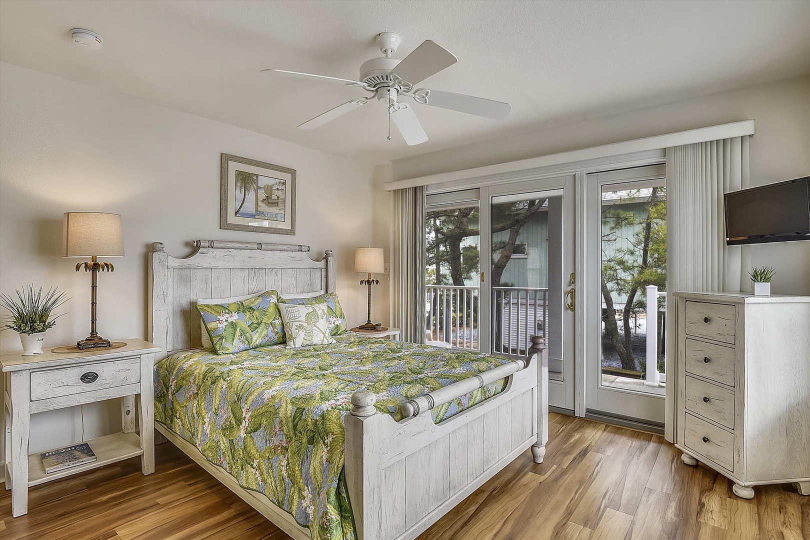 Your coastal retreat awaits with a breezy ceiling fan, whitewashed furnishings, and tropical-themed bedding. Step through glass doors to your private balcony framed by natural greenery.