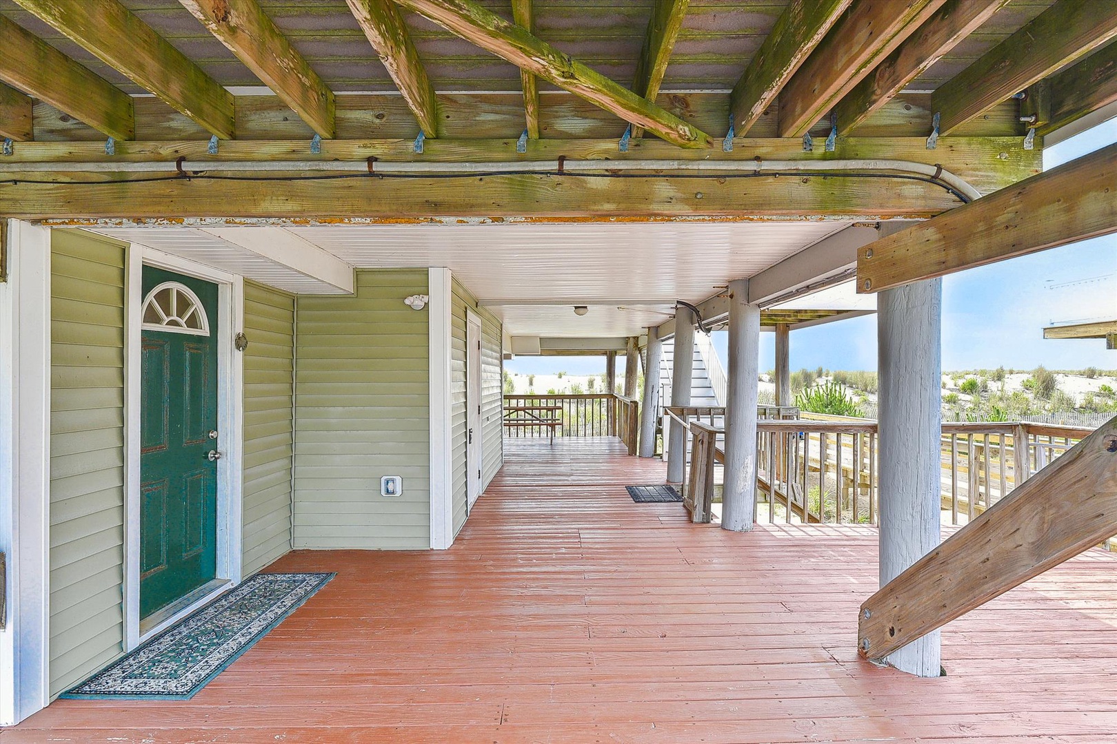 A welcoming beachfront entrance with a wraparound porch featuring rustic timber beams and coastal dune views beyond the railing.