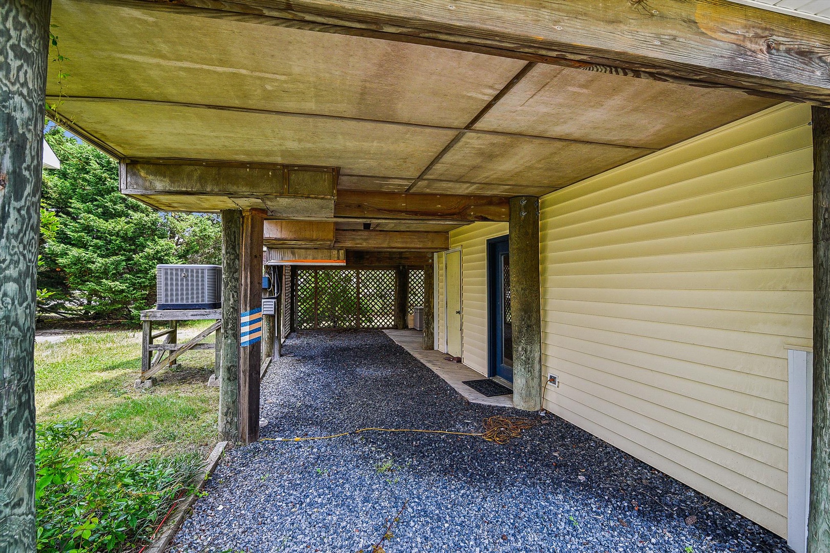 Covered carport with gravel flooring, wood-beam construction, and lattice screening, adjacent to property entrance and natural wooded surroundings.