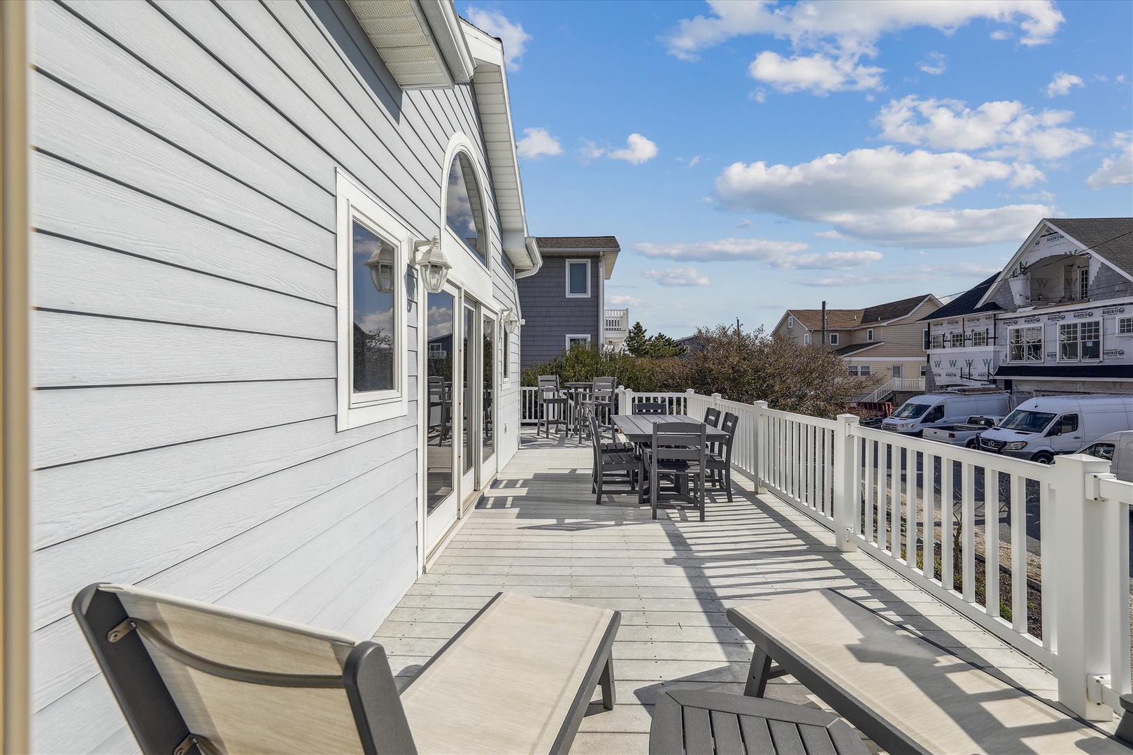 Spacious deck with lounge chairs and dining setup, perfect for sunny breakfasts and evening relaxation. Coastal neighborhood views and open skies frame this inviting outdoor space.