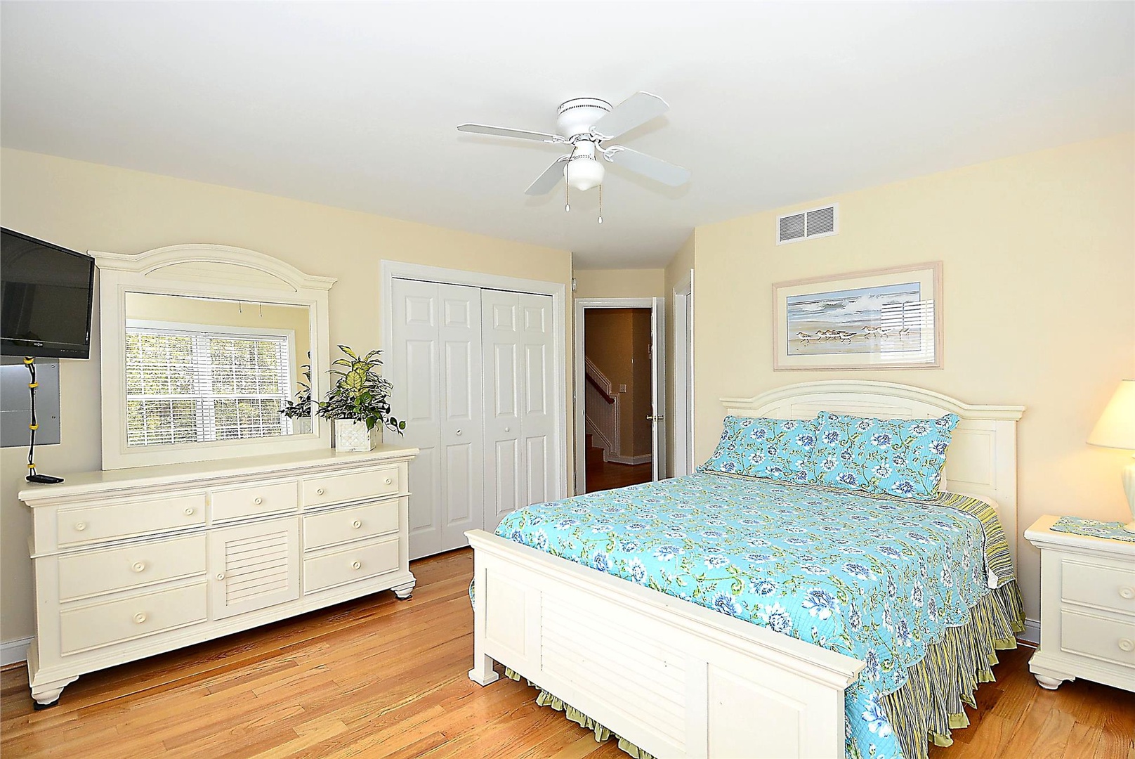 Your inviting bedroom features elegant white furniture, a ceiling fan for comfort, hardwood floors, and cheerful coastal bedding—perfect for peaceful rest after beach adventures.