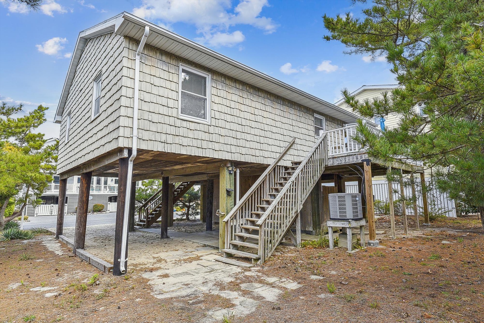 Classic coastal cottage elevated on sturdy pilings, featuring a private entrance stairway and spacious covered parking, nestled among mature pine trees in a quiet beach community.