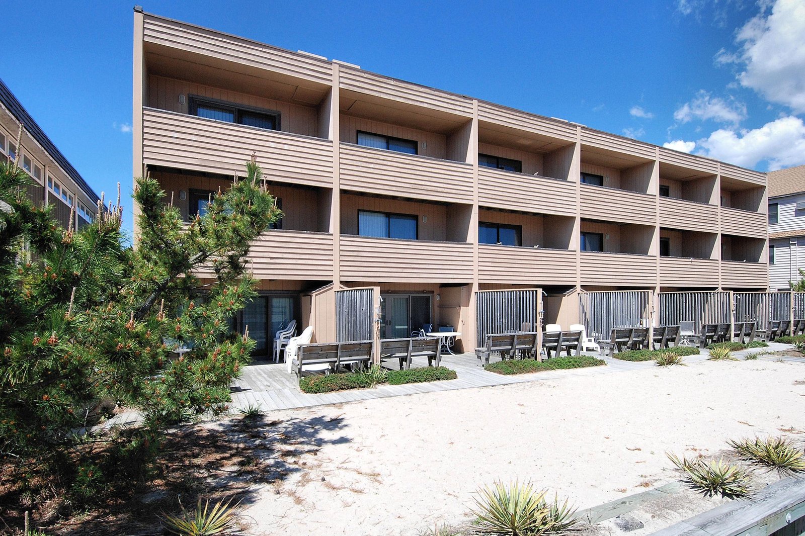 A beachfront building with ground-floor balconies and private seating areas, positioned directly on the sand under summer skies.