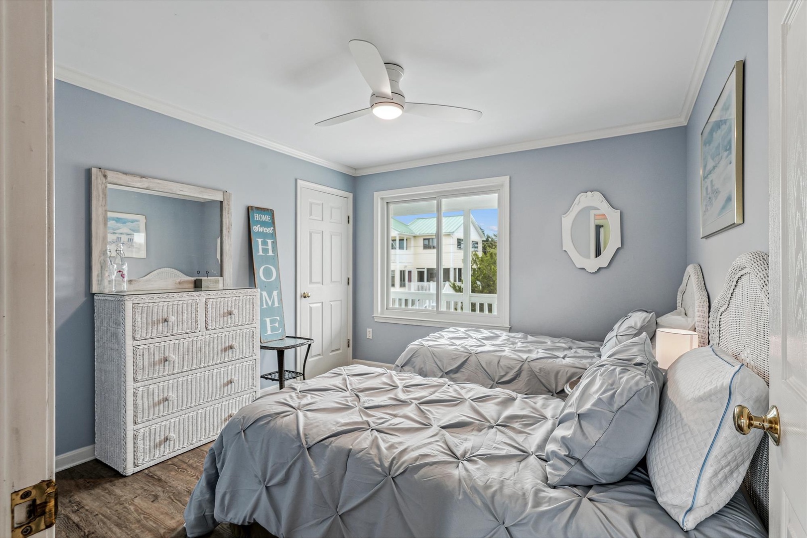 This serene bedroom features calming coastal blue tones, two comfortable beds with soft linens, a ceiling fan to keep you cool, and a window letting natural light stream in, creating your perfect beach retreat.
