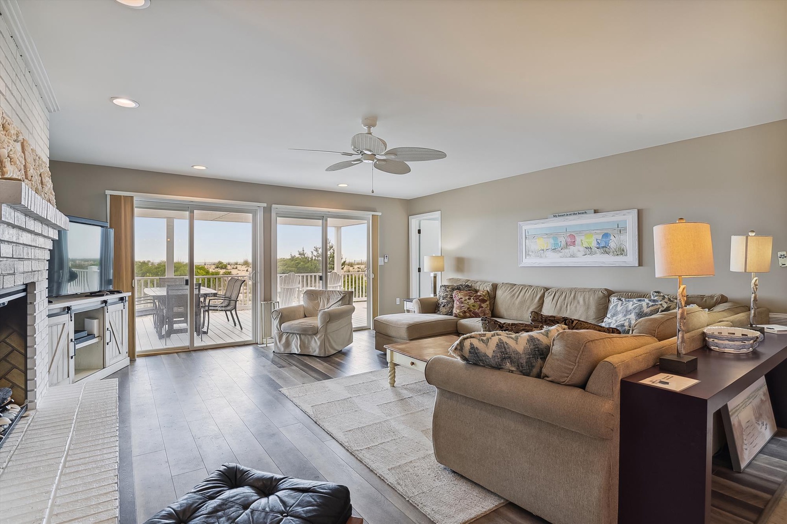 Step into your spacious living room where natural light floods through floor-to-ceiling glass doors, revealing your private balcony. Sink into plush sofas, enjoy the coastal breeze from the ceiling fan, and unwind by the warm fireplace after beach days.