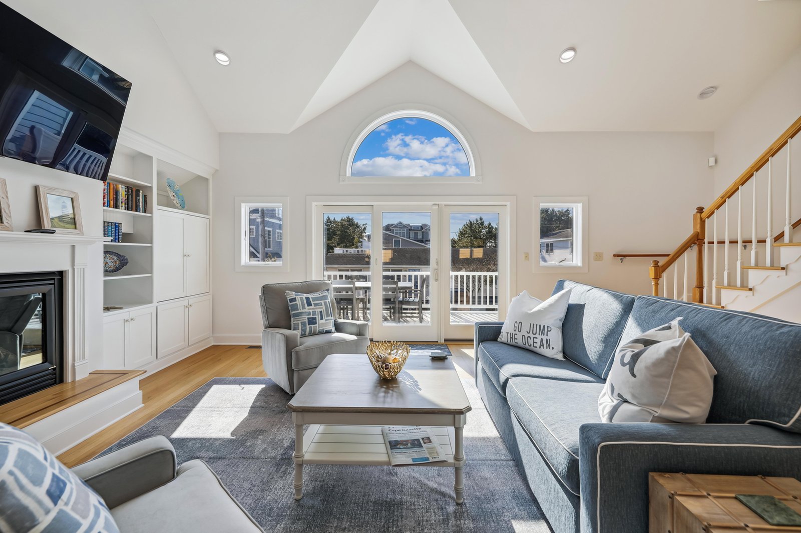 This light-filled living room welcomes you with soaring vaulted ceilings and a stunning arched window that frames the sky. Sink into the plush sofa by the fireplace, soak in the coastal charm, and step out through French doors to your private deck.