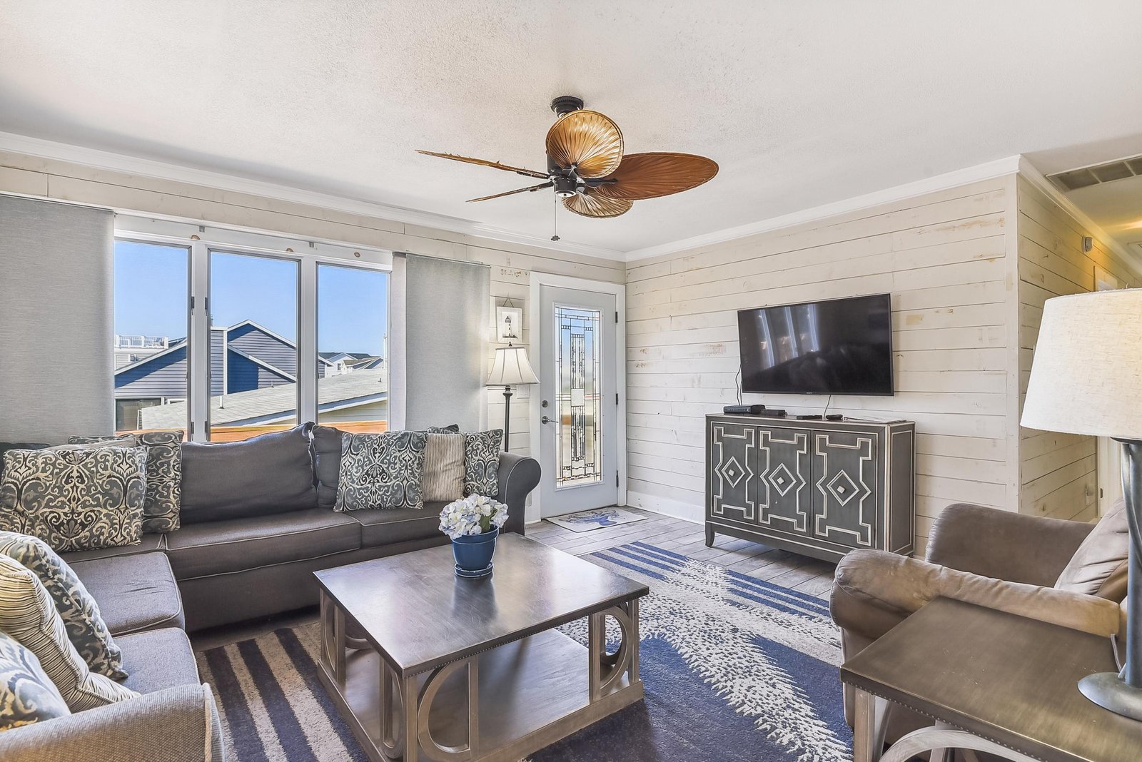 Step into your bright coastal living space, where shiplap walls and a tropical ceiling fan set the tone for relaxation. Large sliding doors flood the room with natural light, opening to your own private outdoor retreat.