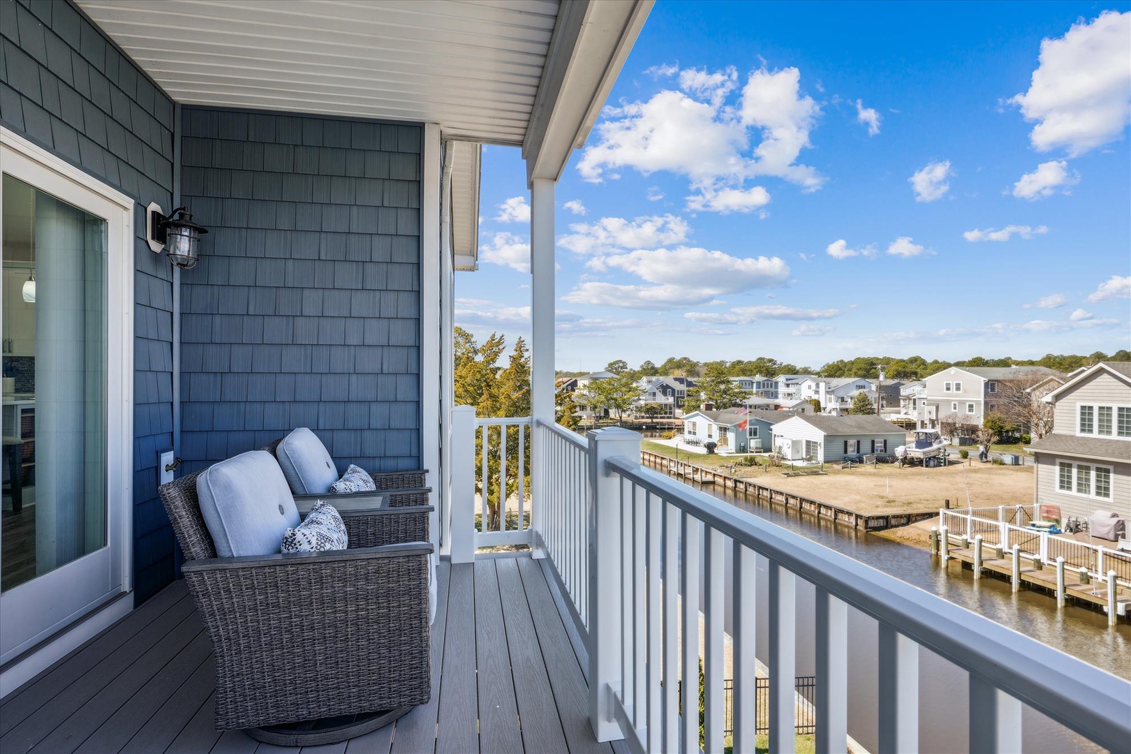 Coastal charm with waterfront community views. Relax on the private balcony and watch boats glide by from comfortable wicker seating under a bright afternoon sky.