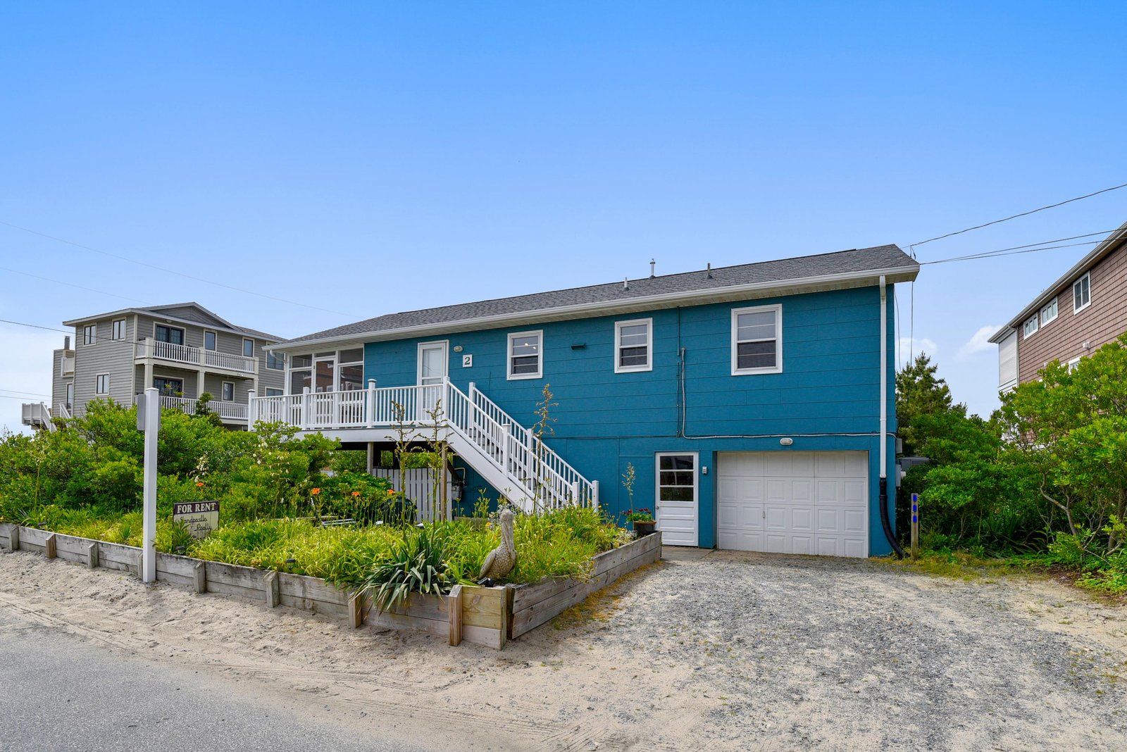 A bright blue beach house on stilts with stairs leading up to a spacious deck, nestled in a coastal neighborhood with sandy surroundings and lush greenery.