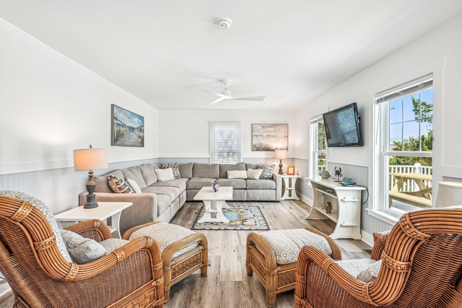 Unwind in your bright coastal living room with plush seating and cool ceiling fans, where natural light pours through generous windows overlooking lush greenery.