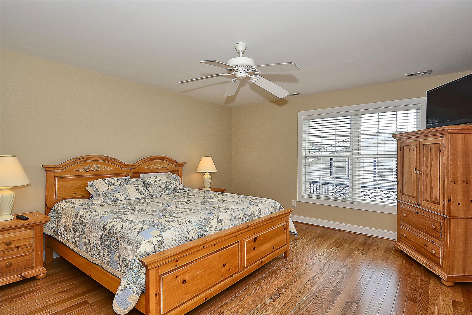 Your spacious bedroom features warm wood furnishings, a ceiling fan to keep you comfortable, and natural light streaming through large windows overlooking the neighborhood.