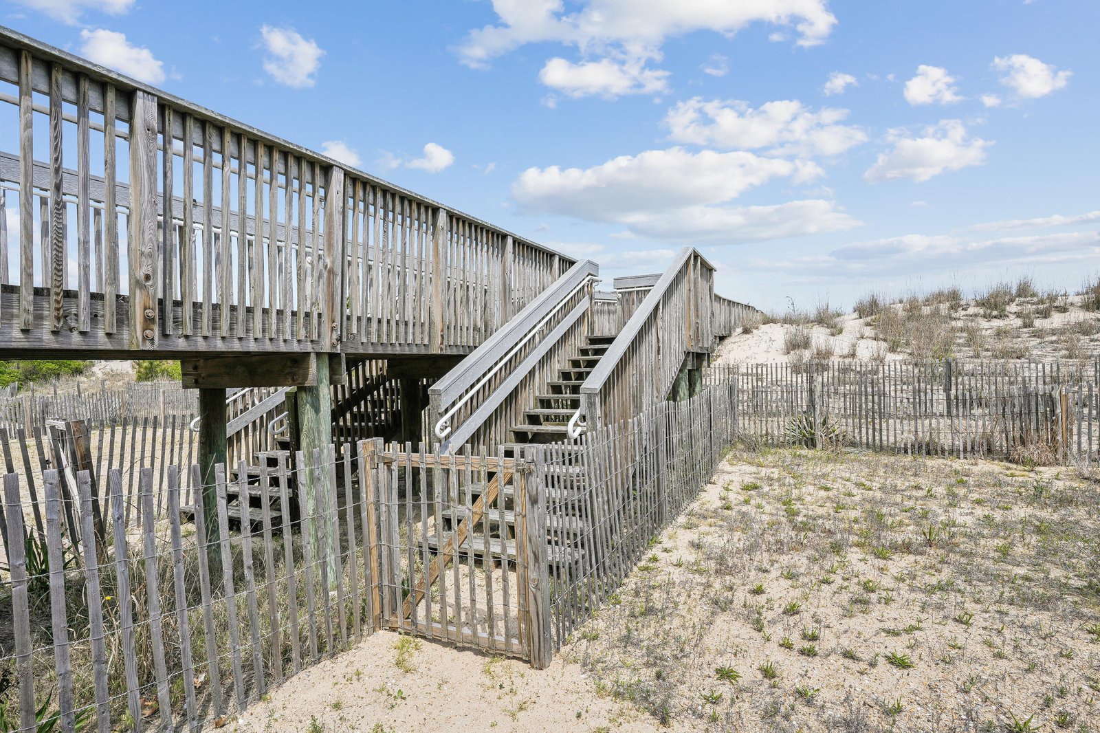 Weathered wooden boardwalk leading over sandy dunes toward the beach, with coastal fencing and native grasses dotting the path—your gateway to oceanside relaxation.