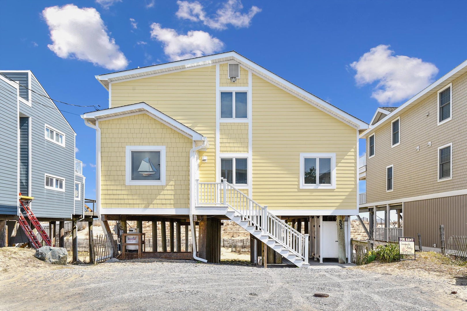 A cheerful yellow coastal home elevated on stilts, situated in a beach neighborhood with neighboring properties on either side under bright blue skies.