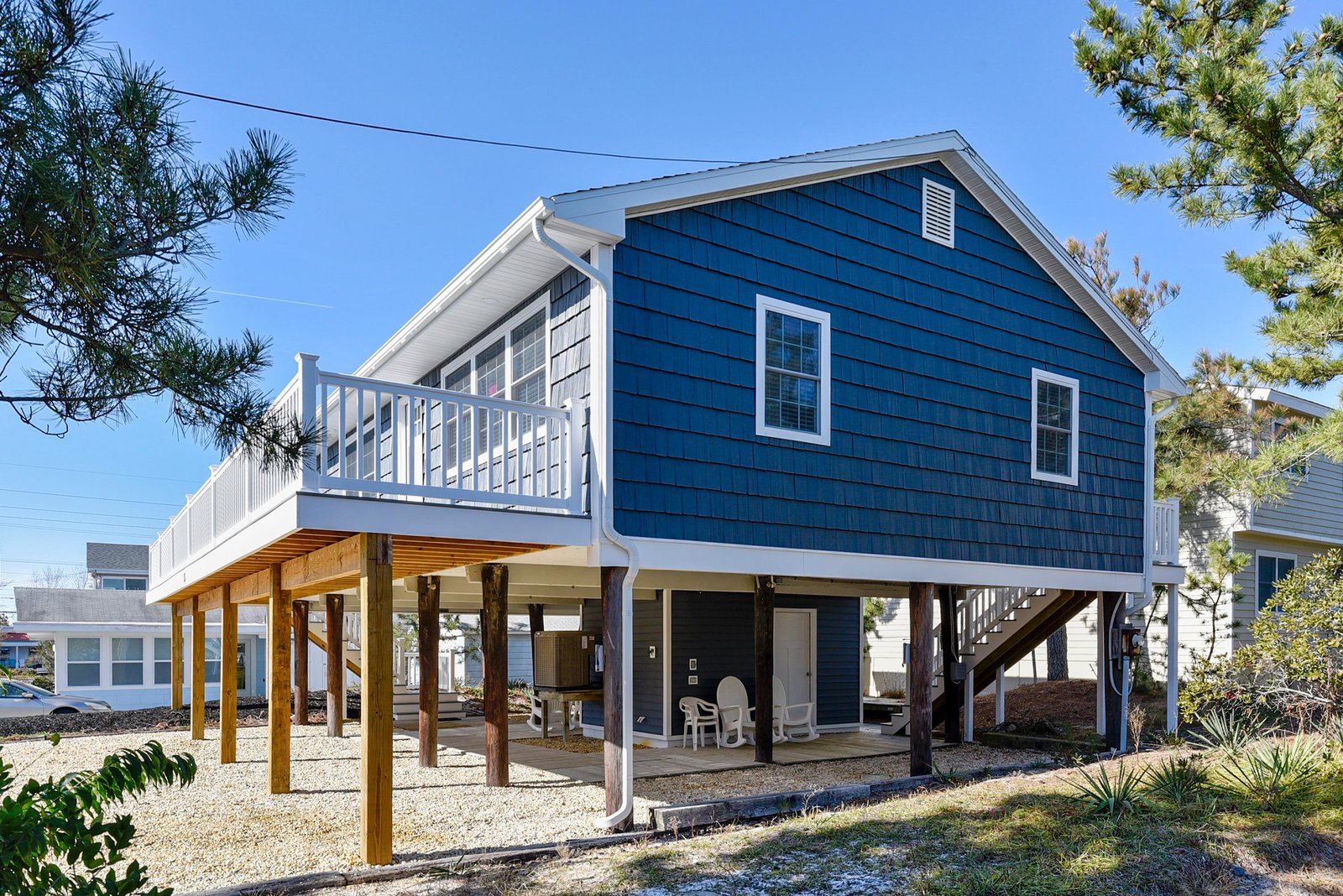 A charming coastal beach house elevated on sturdy pilings, featuring crisp blue siding, white trim, and an inviting upper-level balcony overlooking the surrounding area.