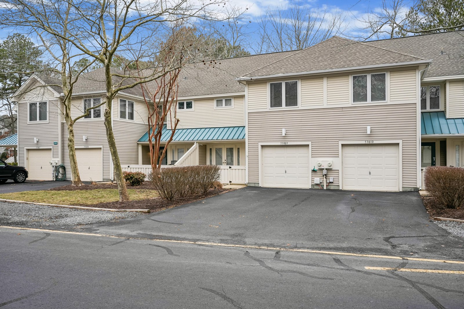 Two-story townhome-style building with beige siding and turquoise metal awnings located in a residential community. Features attached garages and landscaping with deciduous trees and shrubs, photographed in late winter or early spring.