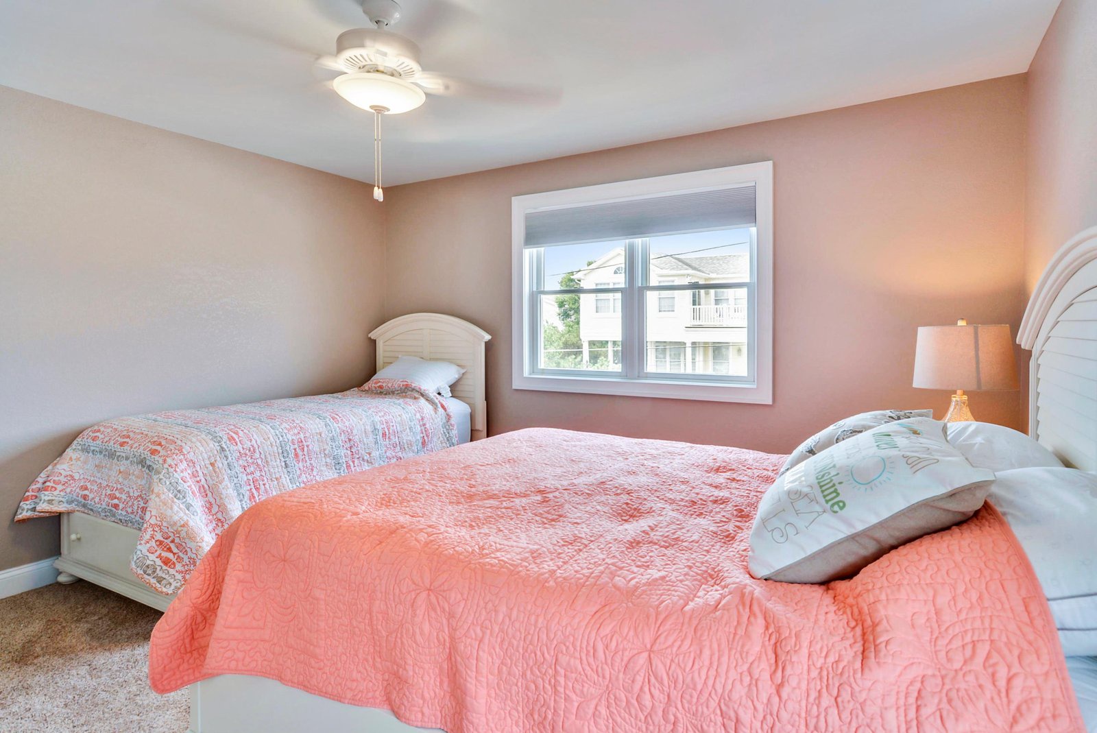 Two cozy beds await, dressed in soft coral and peach quilts that invite you to unwind. Natural light streams through the window while a gentle ceiling fan keeps your room comfortable.