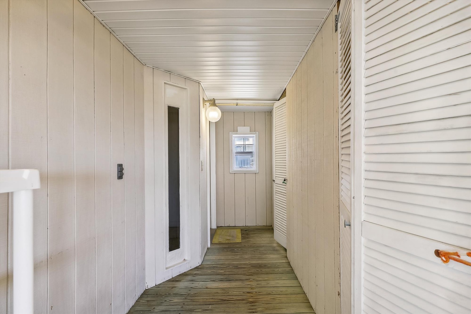 A bright, well-lit hallway connects rooms throughout the home, featuring classic wood paneling and natural light from the window ahead. The warm wood floors and louvered doors add coastal charm.