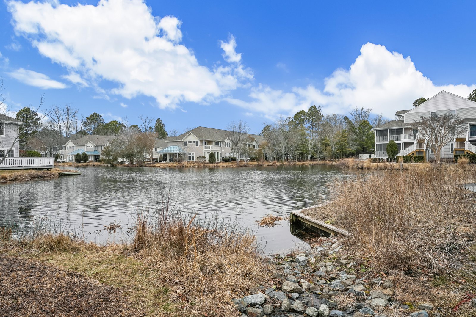 A tranquil pond community setting with private dock access, surrounded by natural marshland and neighboring homes under expansive blue skies.
