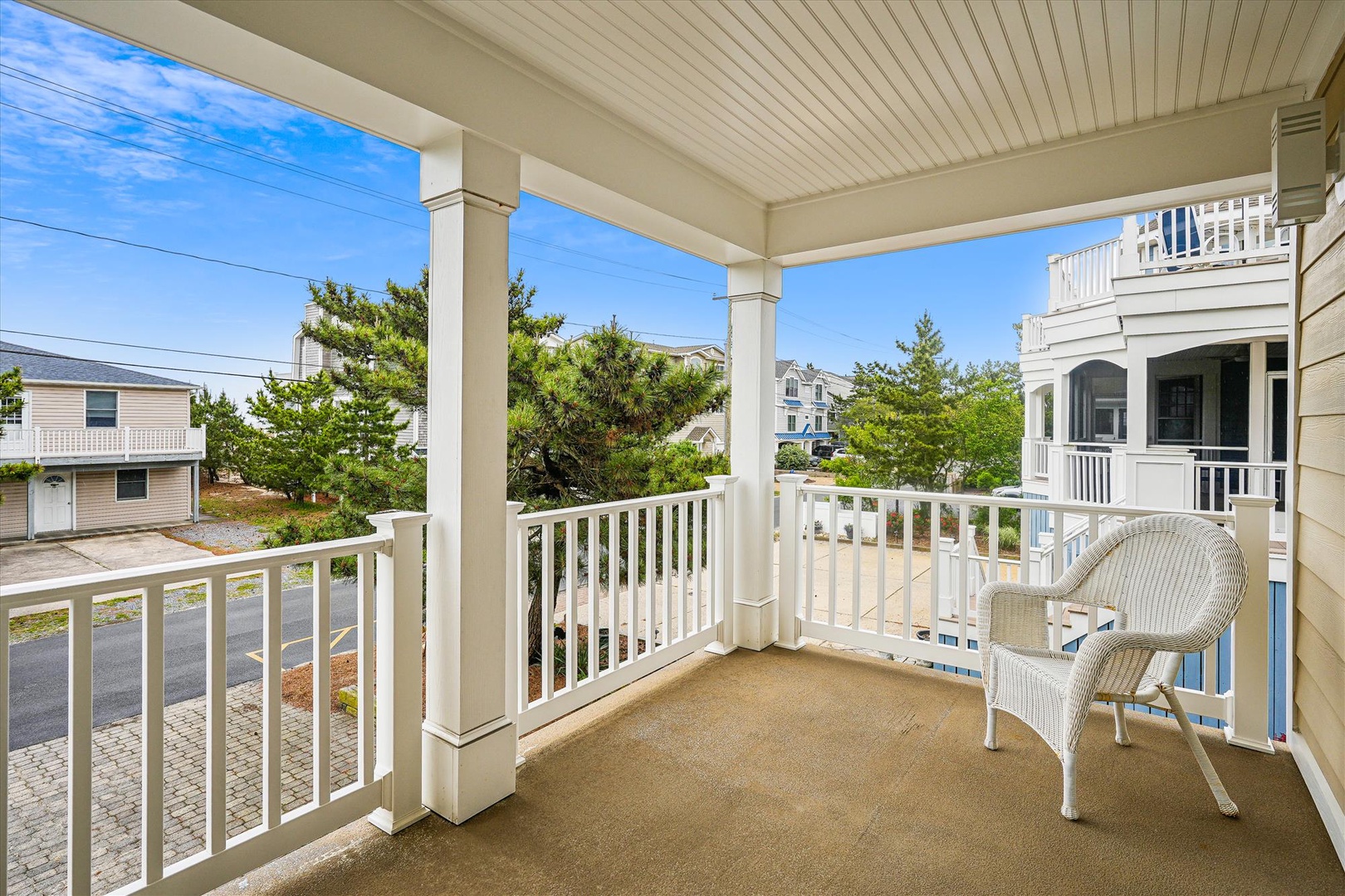 A covered balcony overlooks a quiet residential street, framed by neighboring beach homes and lush greenery. White wicker seating invites you to relax under the beadboard ceiling and enjoy the coastal breeze.