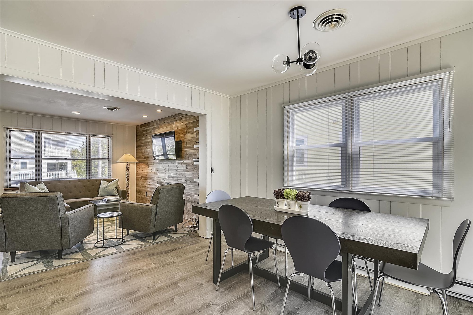 Gather around the farmhouse-style dining table as morning light streams through tall windows. Relax into the cozy living area beyond, where modern charm meets warm wood accents and plush seating.