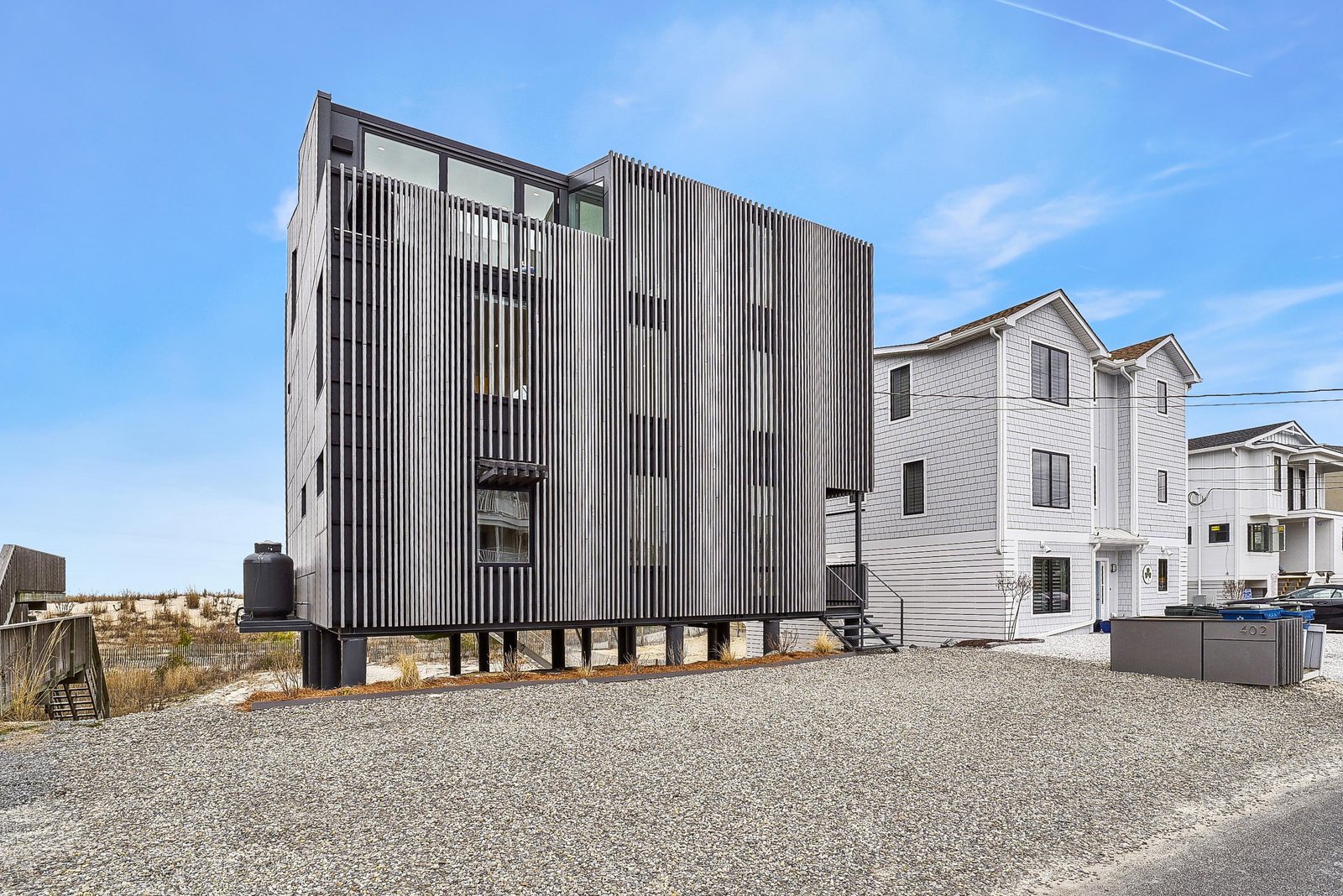 A modern elevated beach home with striking vertical metal siding stands near the coast, surrounded by gravel and neighboring properties under a clear blue sky.